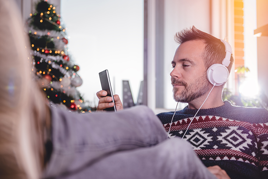 Man wearing blue sweater using smart phone listening to music at home with Christmas tree in background